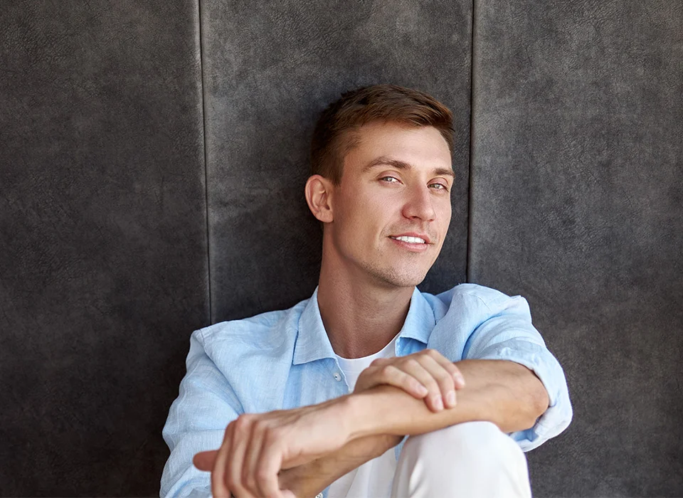 A young man with short brown hair, wearing a light blue shirt, sitting against a dark textured wall, smiling - Dental Exams and Checkups in Beverly Hills, CA