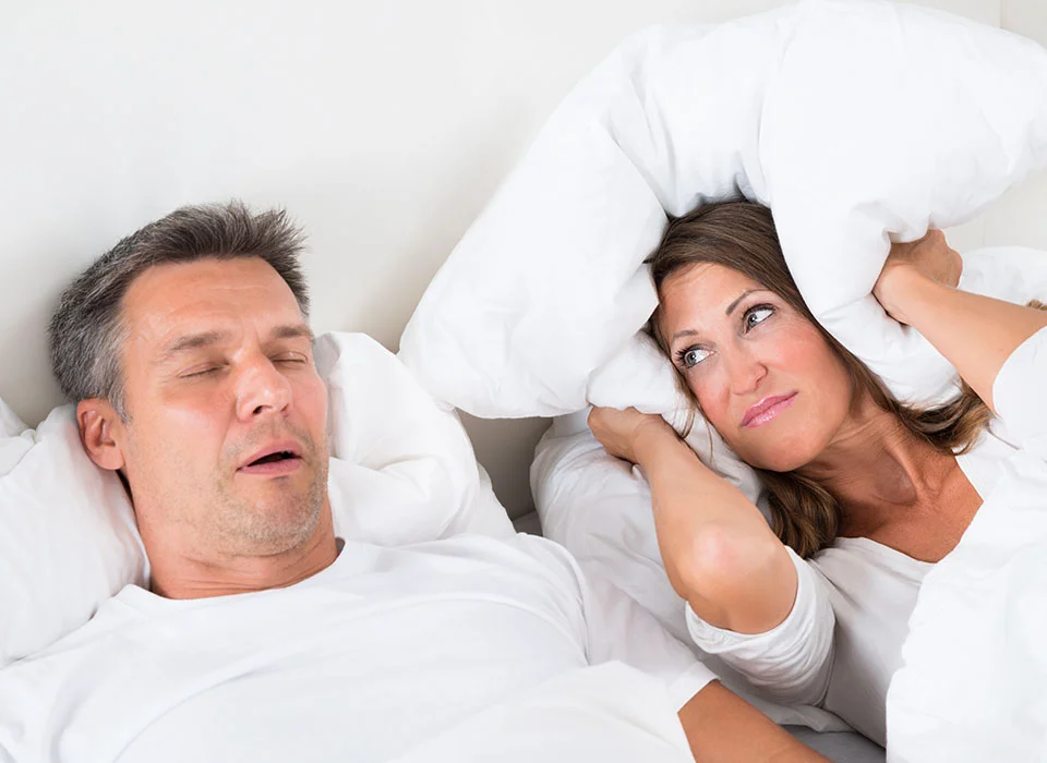 Man snoring while sleeping beside a woman covering her ears with a pillow, indicating the impact of sleep apnea on partners - Sleep Apnea in Beverly Hills, CA